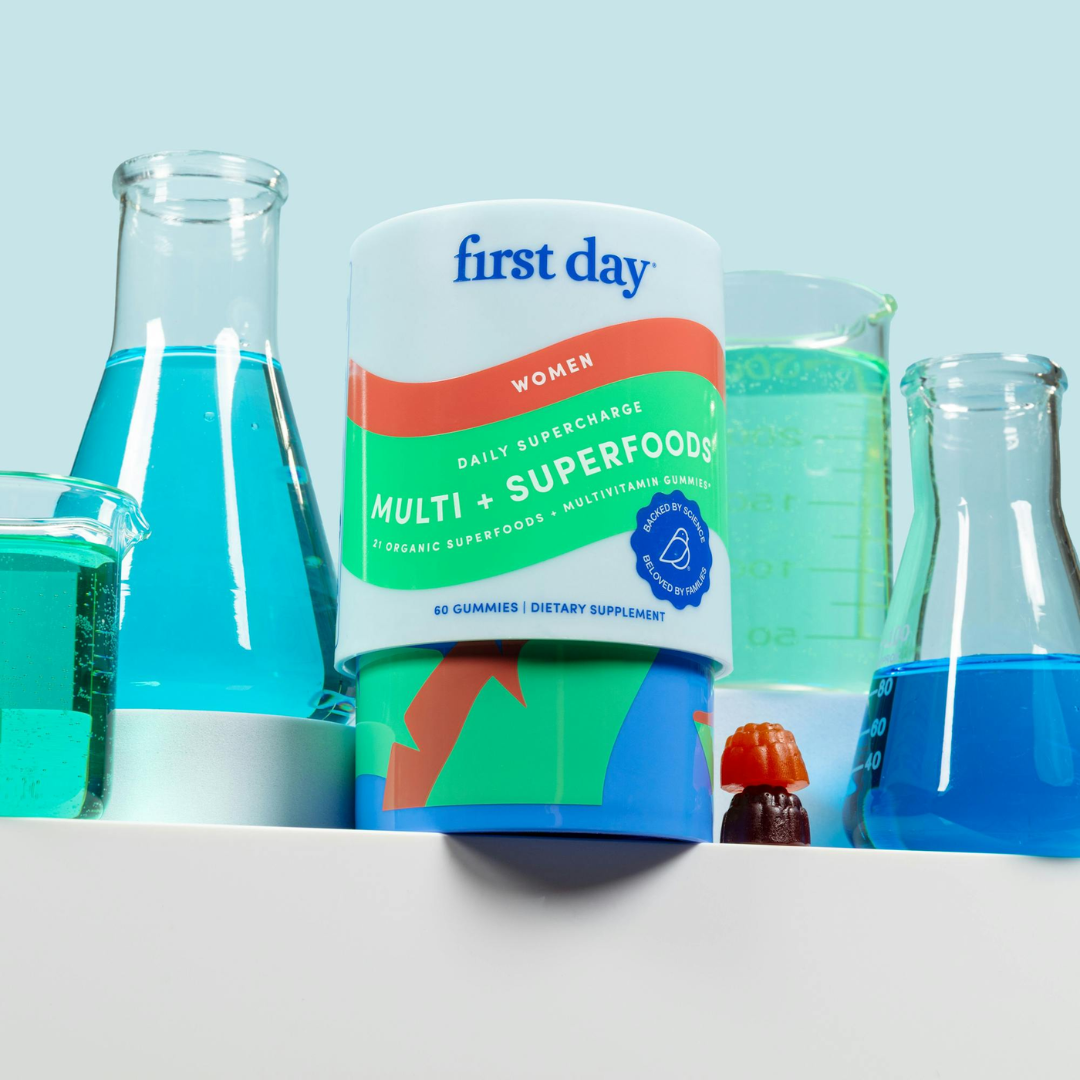 A container of First Day Women’s Multi + Superfoods gummies is displayed on a shelf, surrounded by laboratory glassware filled with blue liquid and a single gummy placed in front of the container.