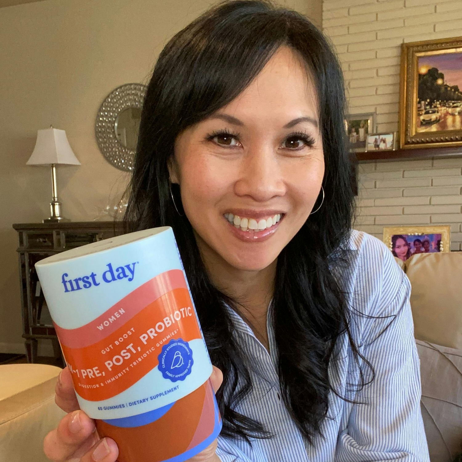 A smiling woman with long dark hair holds up a container labeled first day Pre, Post, Probiotic in a living room with a lamp, couch, and picture frames in the background.
