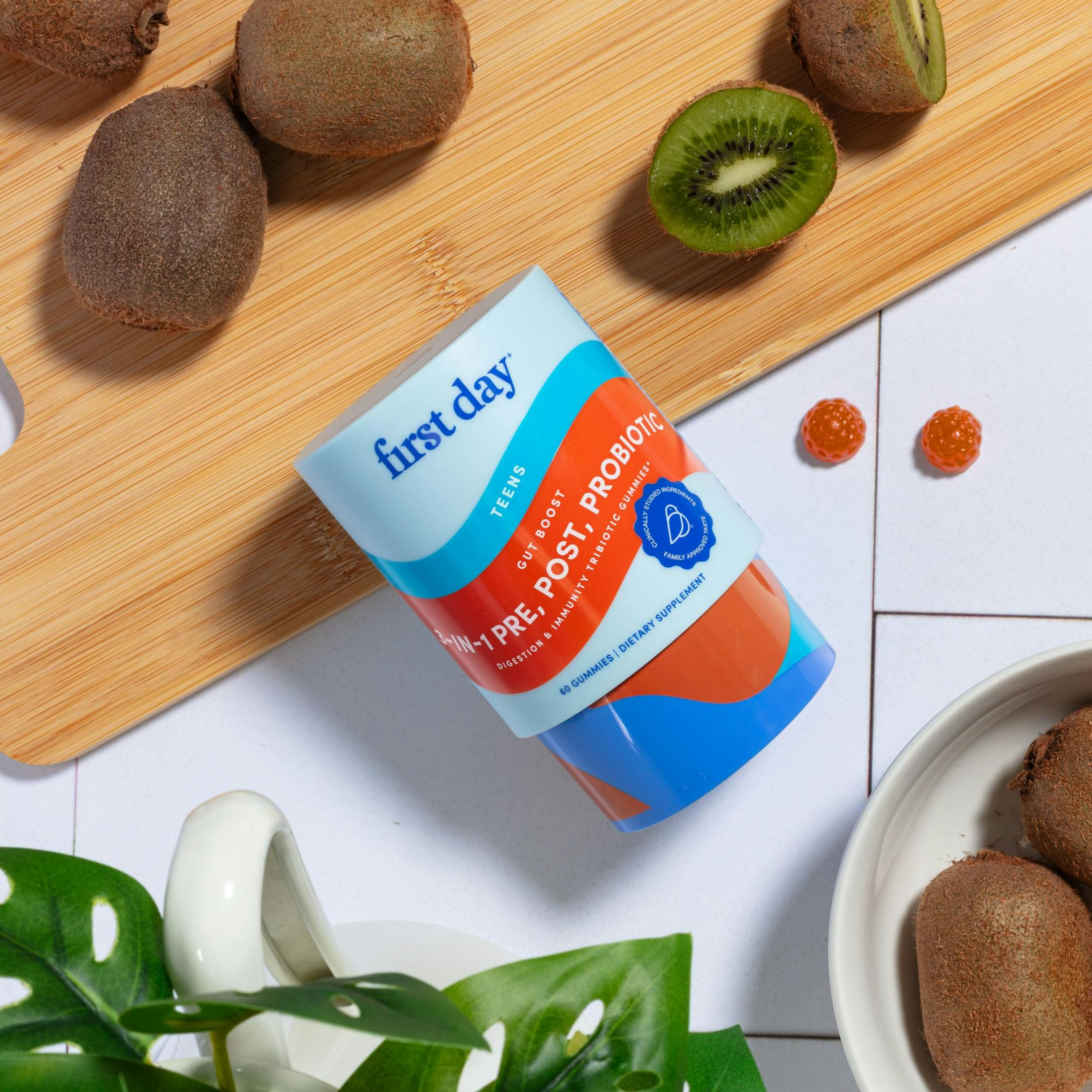 A container of First Day Teen Pre + Post Probiotic is on a white surface beside sliced and whole kiwis on a wooden board, gummy supplements, and a leafy green plant.