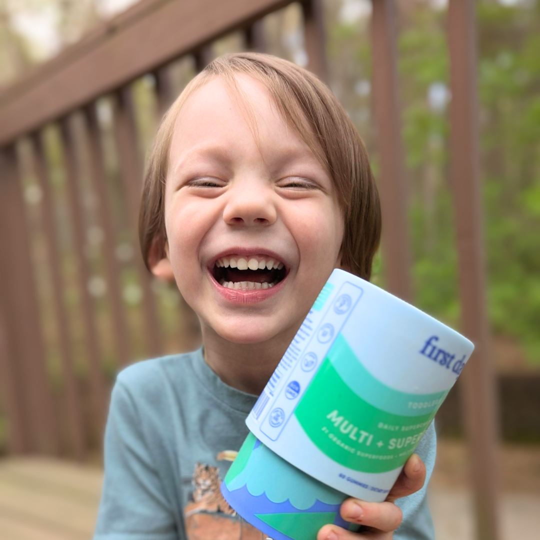 A young child with light brown hair is smiling widely and holding a container of toddler multivitamin powder while standing outside on a wooden deck. The background is blurred with green foliage visible.