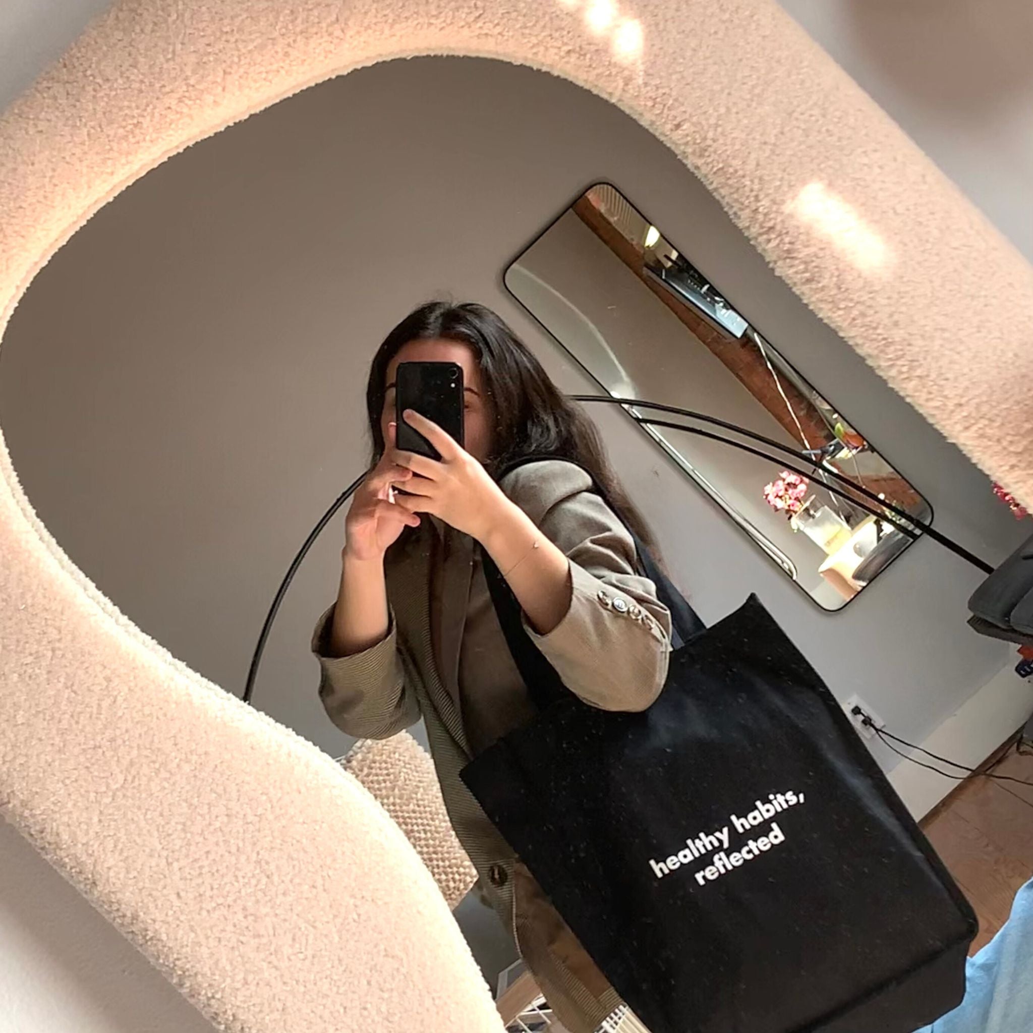 In a cozy room, someone takes a mirror selfie holding the Reflection Tote, which reads "healthy habits." The mirror’s wavy, textured frame is eye-catching, with another mirror and flowers visible in the background.