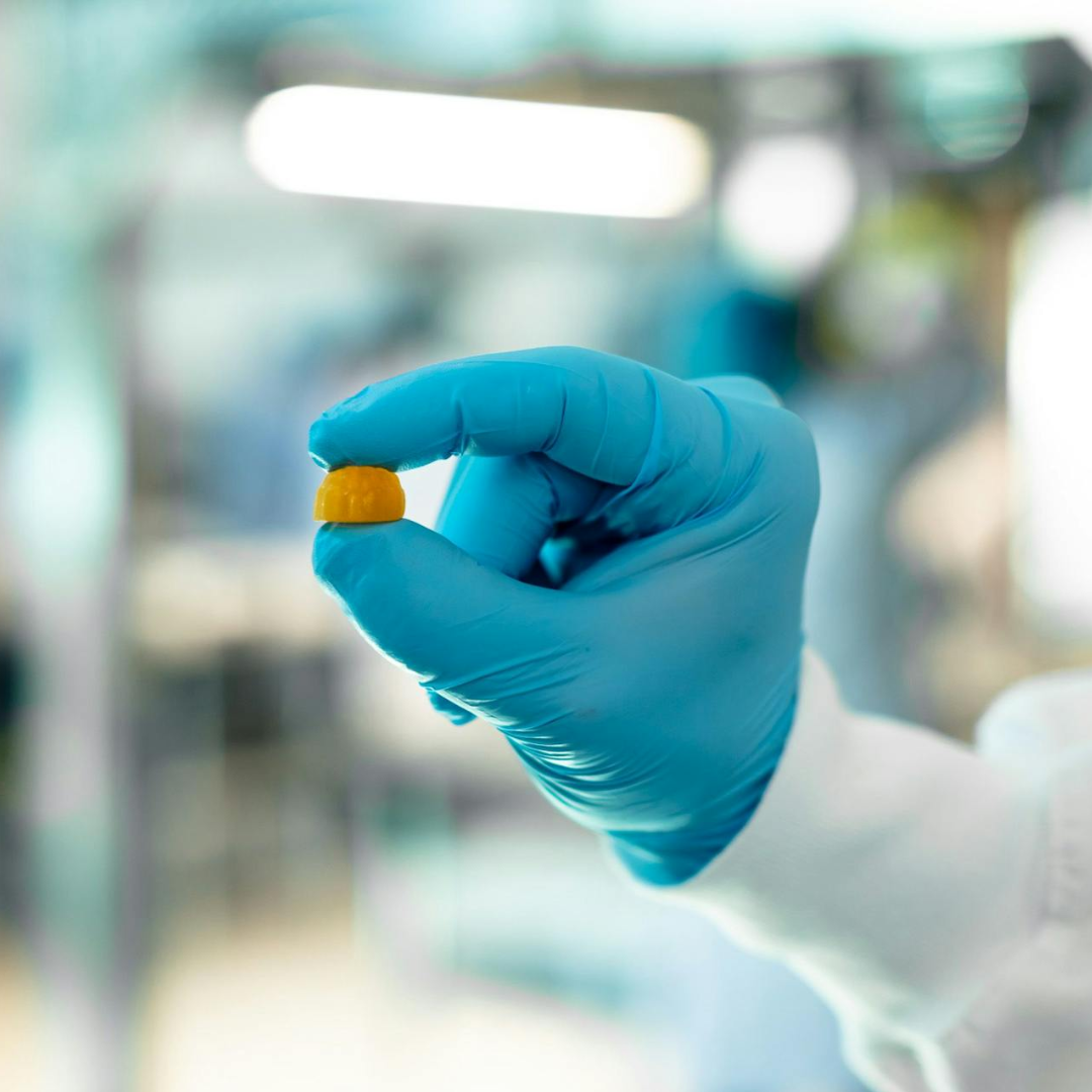 A person wearing a blue glove holds a small yellow pill or tablet between their fingers in a laboratory setting. The background is blurred, focusing on the gloved hand and the pill.