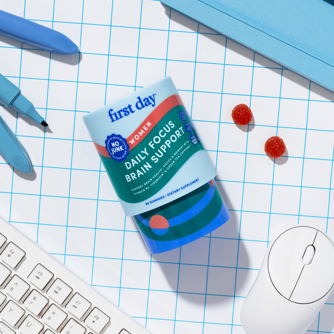 Packaging of 'first day' women's daily focus brain support supplement on a grid background with pens and a computer mouse.