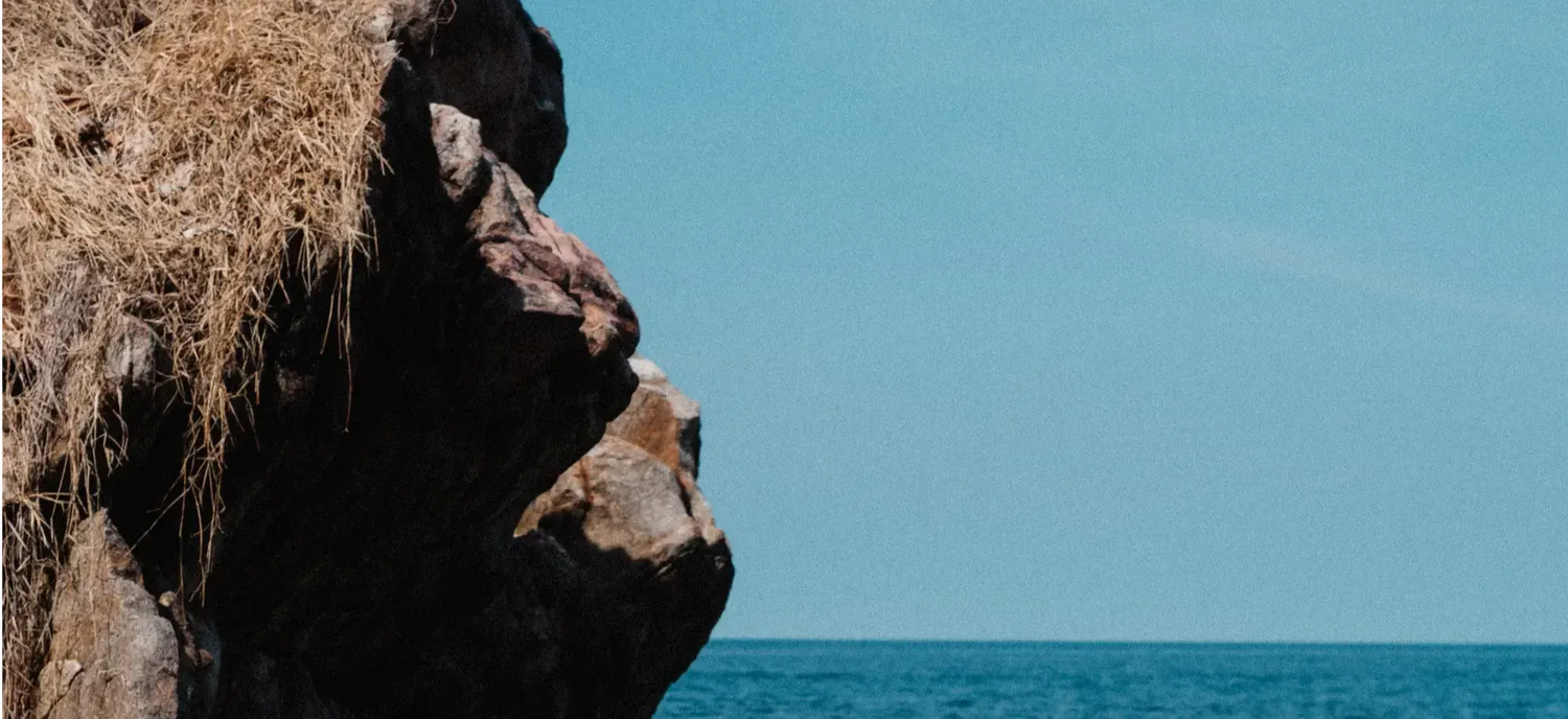 A rocky cliff with dry grass on the left overlooks a calm blue sea under a clear sky.
