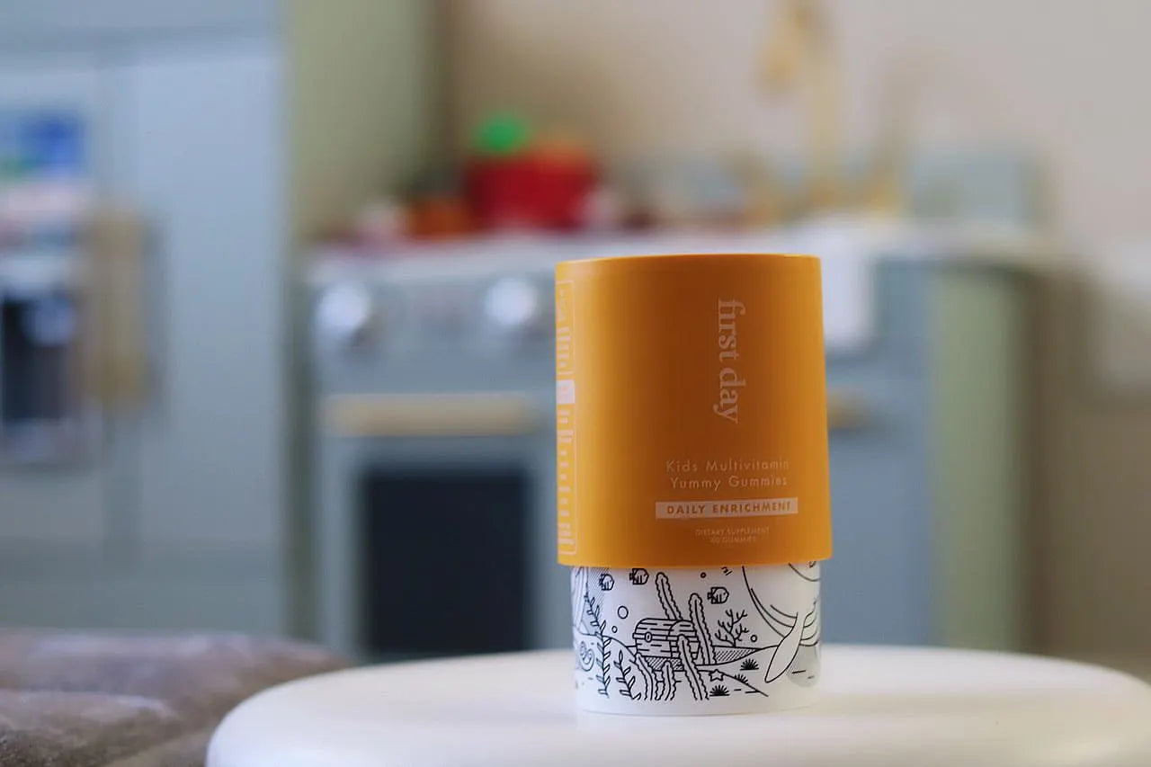 A container of First Day Kids Multivitamin Yummy Gummies stands on a table, with a yellow label on top and a white base featuring black line art. A blurred kitchen is visible in the background.