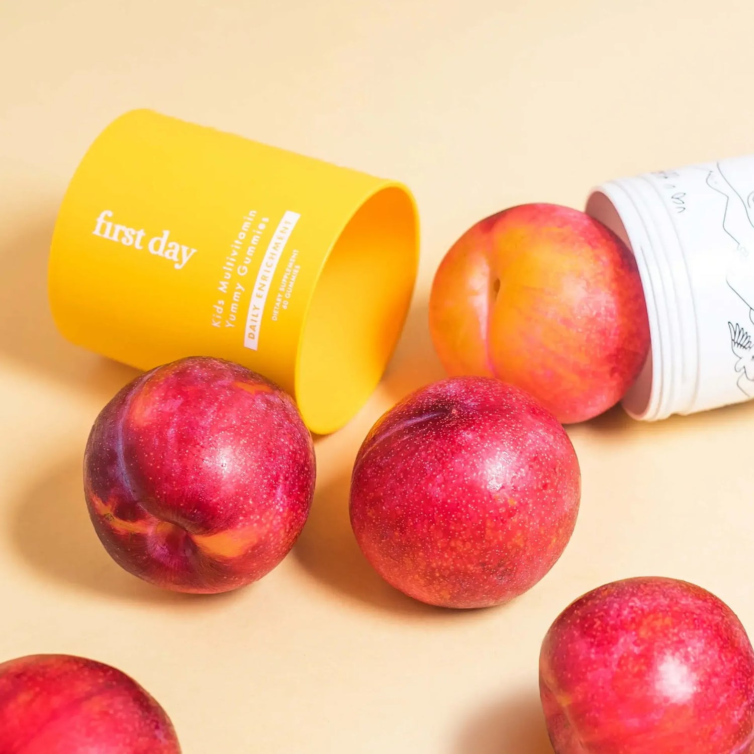 Two colorful containers, one yellow and one white, lie on their sides, with round, red and yellow plums spilling out onto a light beige surface. The containers are labeled first day and feature playful designs.