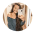 A young woman with long brown hair smiles and takes a mirror selfie with her phone, wearing an off-the-shoulder blue top, standing in a hallway with warm lighting.