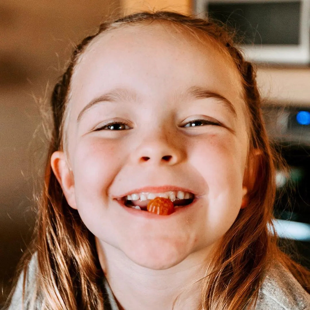 A young girl with long light brown hair smiles widely, showing a missing tooth and a small gummy candy in her mouth. She is indoors with warm lighting.