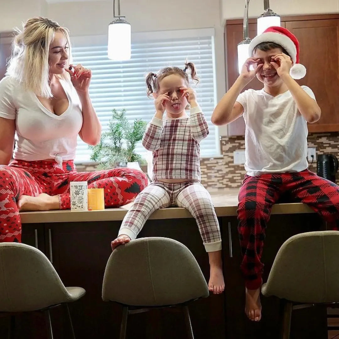 A woman and two children sit on a kitchen counter in festive pajamas. The children playfully hold their fingers in circles over their eyes, and one wears a Santa hat. They are smiling and enjoying time together.