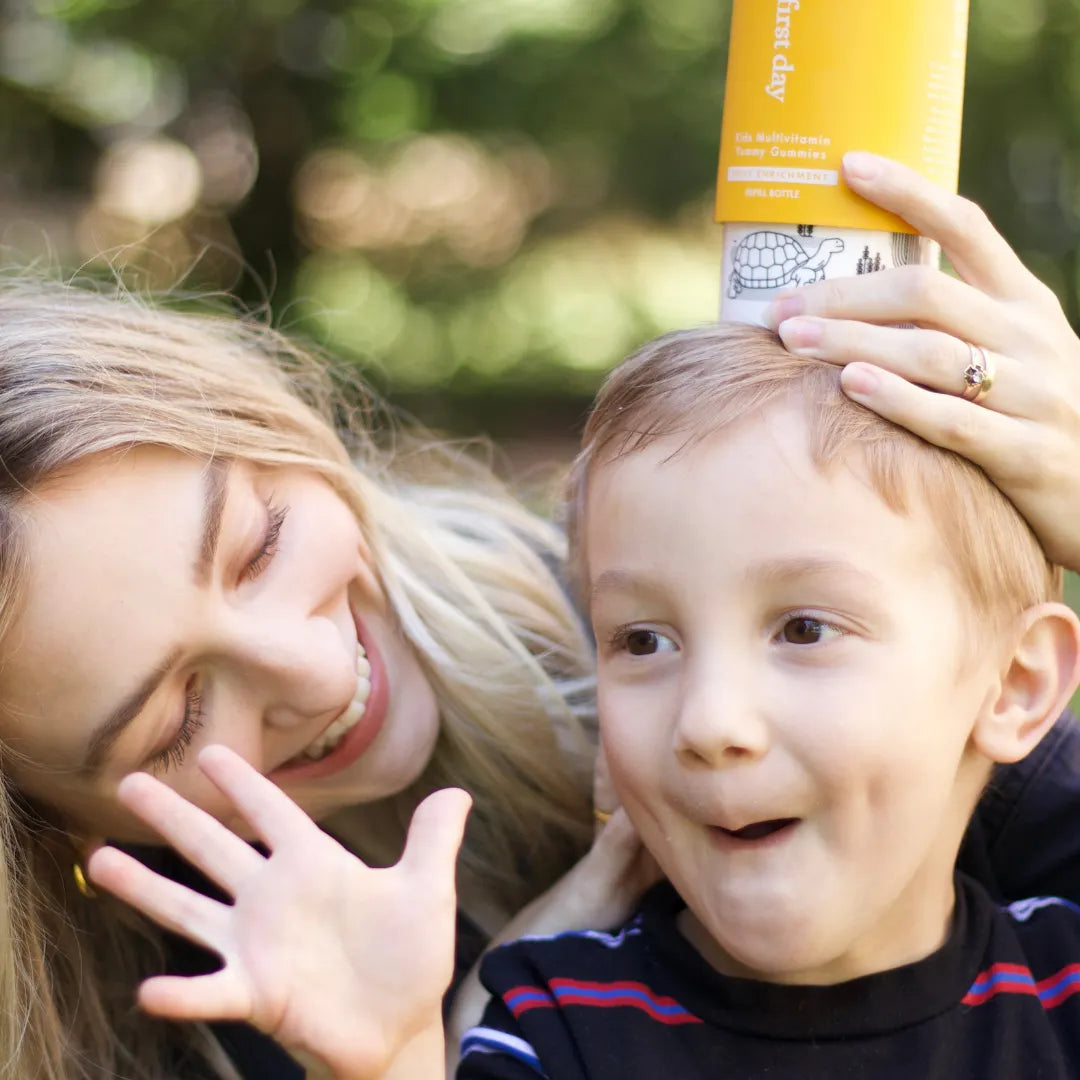 A smiling woman holds a yellow bottle on a young boy’s head while he laughs and waves his hand. They are outdoors, and both look happy and playful.