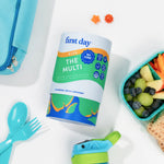 A First Day Kids 'The Multi' dietary supplement canister is displayed in a styled flat-lay on a white surface, surrounded by school and snack props including a blue lunch bag, utensils, and a fruit container.