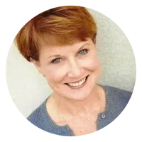 A woman with short red hair, wearing a gray top, is smiling at the camera. The background is light and neutral. The photo is cropped in a circular frame.