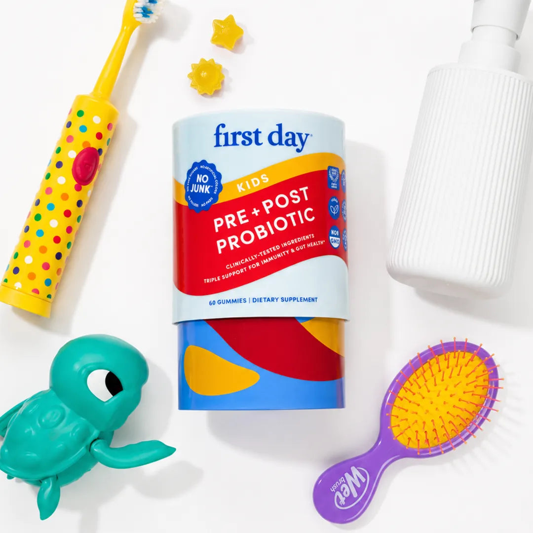 A cylindrical First Day Kids Pre + Post Probiotic container is centered on a white background, surrounded by colorful children's accessories including a yellow polka-dot toothbrush, a teal turtle toy, a purple hairbrush, and a white pump bottle.