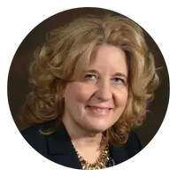 Portrait of a woman with shoulder-length curly blonde hair, smiling, wearing a dark blazer and a gold necklace, shown against a dark background inside a circular frame.