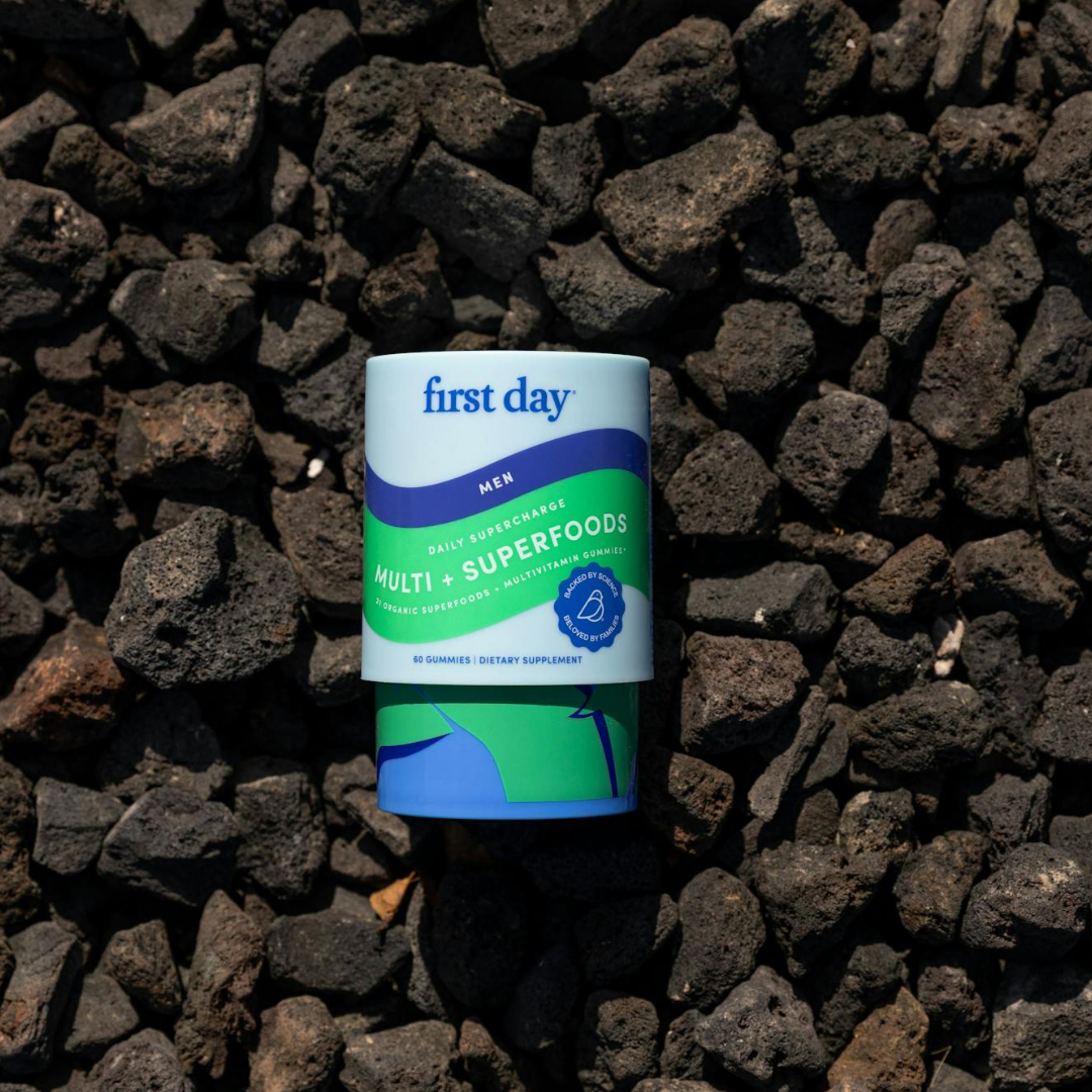 A container of First Day Mens Multi + Superfoods dietary supplement rests on a bed of dark, rough volcanic rocks. The container is green and blue with white accents and contains 60 gummies.