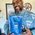 A smiling man with glasses and a beard holds a blue bag of First Day Mens Daily Provisions Multi Gummy Drops. He wears a blue sweatshirt with white and red writing. The background shows a plant and home decor.