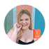A smiling woman with long blonde hair holds an orange mug and appears to be eating chocolate. She stands in front of a colorful wall with green, orange, and blue vertical stripes.
