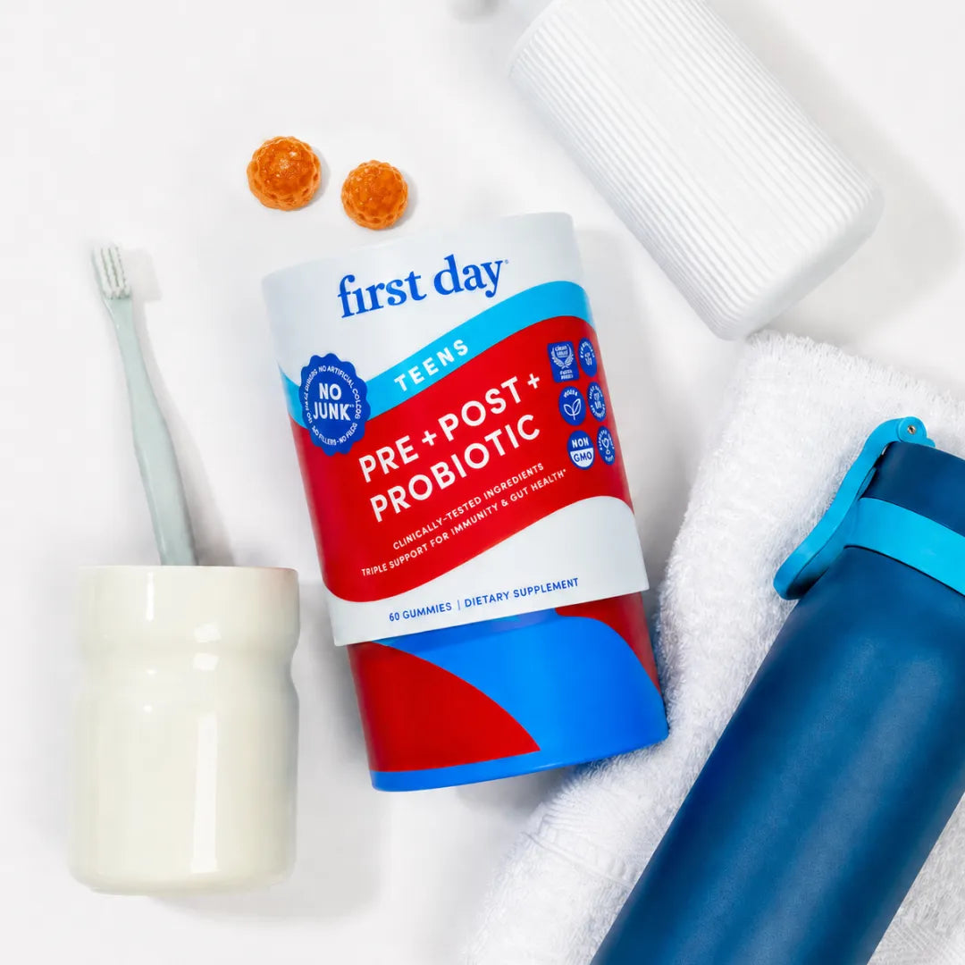 A First Day Tees Pre &amp; Post &amp; Probiotic supplement bottle is displayed in a flat-lay arrangement with dental and hydration props, including a toothbrush, toothpaste tube, toothpick holder, and water bottle, on a white background.