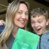 A smiling woman and an excited boy hold a green package labeled “first day Teen’s Daily Kickstart” while posing for a selfie indoors.
