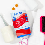 A bottle of First Day Women Pre & Post Probiotic gummies is styled on a white background with bathroom accessories including a toothbrush, cotton pads, a pink hairbrush, and rubber bands.